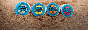 Four tins of Surf Balm on a sandy beach