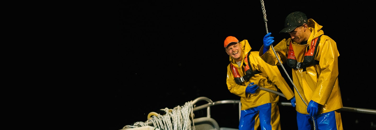 Two men on a fishing boat at night, wearing life jackets and various waterproof garments