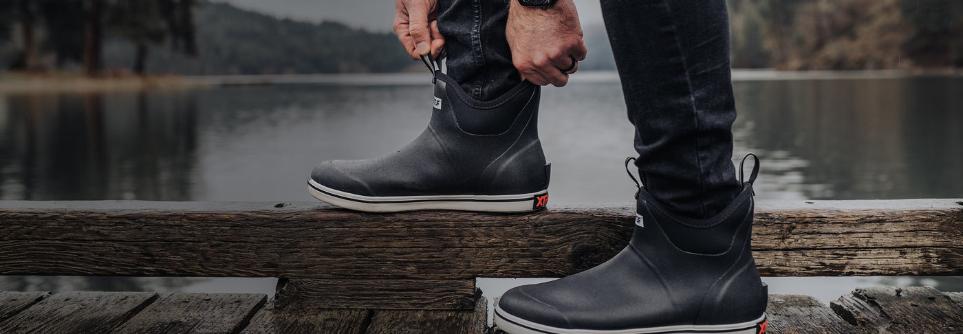 Close-up image of a man putting on a pair of Xtratuf Trolling Pack Fleece Lined Ankle Deck Boots on a wooden jetty over a lake