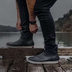 Close-up image of a man putting on a pair of Xtratuf Trolling Pack Fleece Lined Ankle Deck Boots on a wooden jetty over a lake